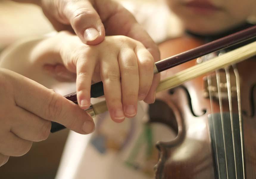 Eine Kinderhand hält einen Geigenbogen und wird von einer größeren Hand gestützt und geführt.