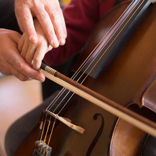 Eine Kinderhand hält einen Cellobogen. Zwei größere Hände korrigieren die Bogenhaltung. Im Hintergrund ist eine kleines Kindercello zu sehen.