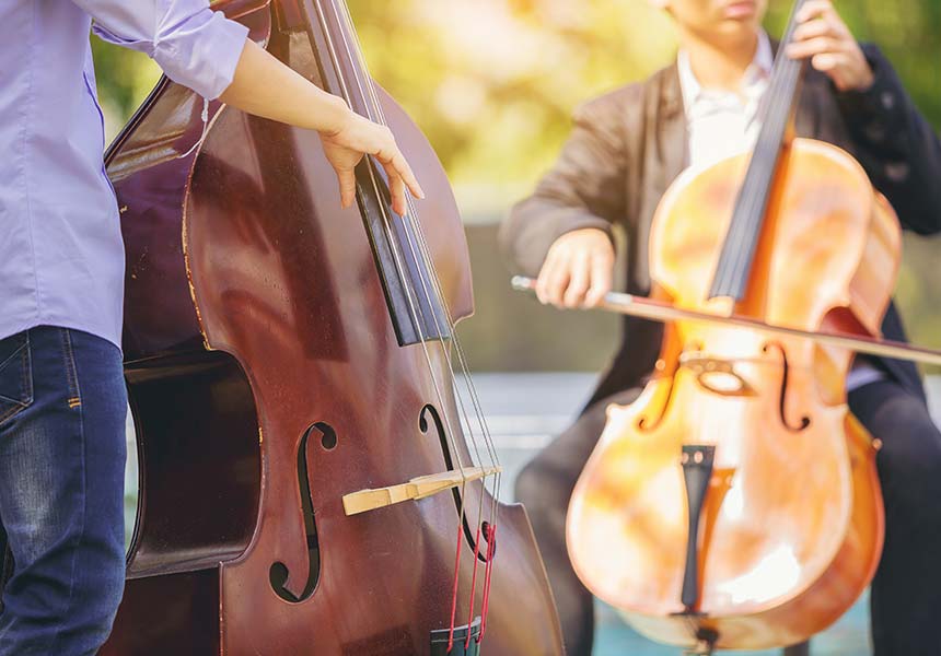 Ein Musiker zupft einem Kontrobass. Der Bass ist dunkelbraun und hat einige Abnutzungsspuren. Im Hintergrund spielt ein anderer Musiker sitzend Cello.