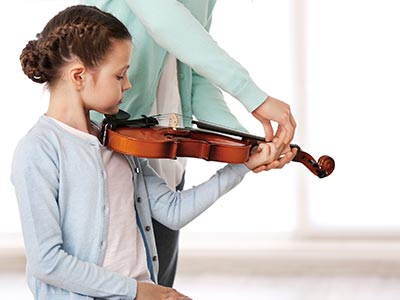 Ein junges Mädchen erlernt das halten der Geige zwischen Kinn und Schulter. Sie sitzt gerade und schaut auf das Griffbrett, ihr Kinn ruht auf dem Kinnhalter und mit der linken Hand hält sie die Geige am Hals fest. Eine größere Person hält die Geige am Griffbrett in Position.